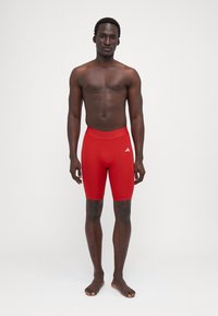Røde atletiske shorts lavet af strækbart stof, tætsiddende design, der når midt på låret, med et hvidt logo på venstre side.