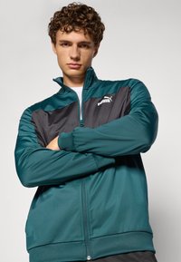 Teal og svart treningsjakke med glidelås i kragen, ribbede ermer og brodert logo på brystet. Slett tekstur, strømlinjeformet design.