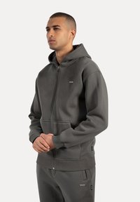 Sudadera gris con cremallera, con un bolsillo tipo canguro, puños y dobladillo acanalados, y etiquetas laterales, hecha de una tela suave y texturizada.
