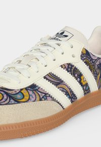Άσπρο sneaker Adidas με πολύχρωμο paisley σχέδιο, τρεις λευκές ρίγες, σκελετό από σουέντ, λευκά κορδόνια και λαστιχένια σόλα.