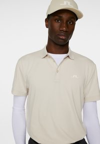 Beige pikétröja med krage och tre knappar, med en vit logotyp på vänster bröst; bärs över en vit långärmad skjorta.