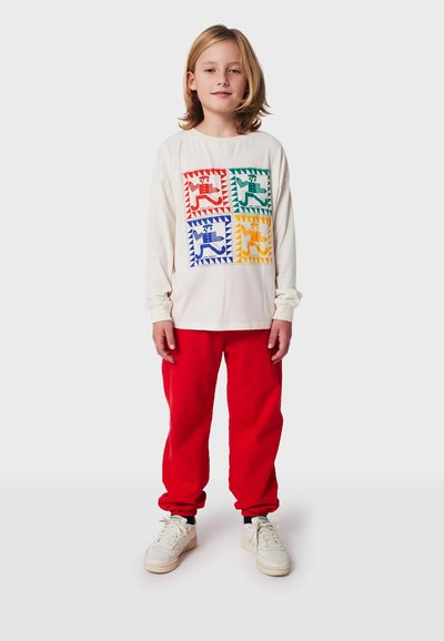Weißes langärmliges Shirt mit bunten geometrischen Tiermotiven, kombiniert mit roten Jogginghosen und weißen Turnschuhen mit grünen Akzenten.