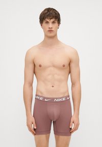 Brūnie Nike bokseru apakšveļa ar stiepjamu jostu un baltu logo, pieguļoša forma un mīksts audums. Nav ievērojamu rakstu vai papildu detaļu.