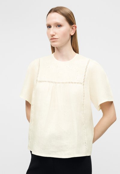 Ξανθιά γυναίκα με ίσια μαλλιά φοράει μια φαρδιά, μπεζ μπλούζα με κοντά μανίκια, διακοσμημένη με κεντημένα σχέδια με τρύπες, και μαύρο παντελόνι.