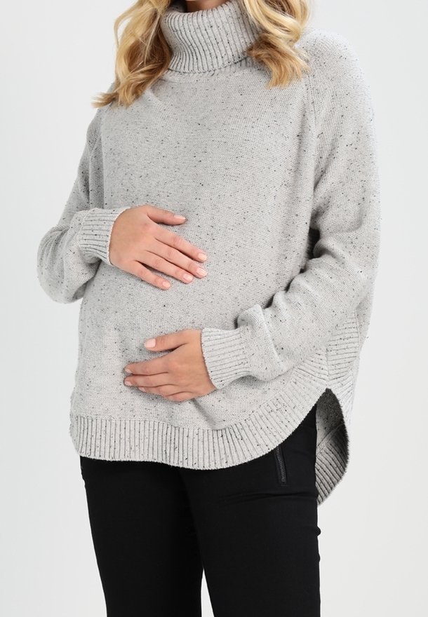 Femme enceinte en pull à col roulé gris tenant son ventre, portant un pantalon noir, debout devant un fond uni.