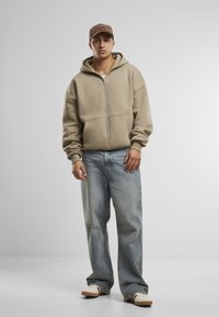 Beige zip-up hoodie met een grote capuchon, gecombineerd met losse blauwe denim jeans en witte sneakers met beige accenten. Bruine pet op het hoofd.