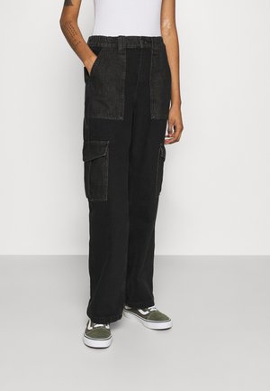 Pantalones cargo de mezclilla negra con dos grandes bolsillos frontales, cinturilla elástica y un corte de pierna ancha. Llevados con zapatillas verdes y una blusa blanca.