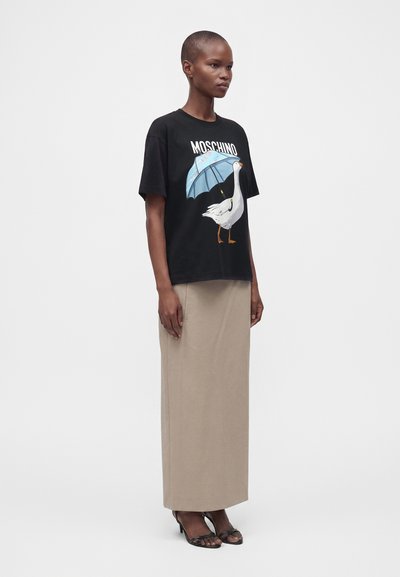 Γυναίκα που φοράει μαύρο t-shirt Moschino με εκτύπωση χήνας και ομπρέλας, μπεζ μακριά φούστα και μαύρες πέδιλες με ανοιχτά δάχτυλα, στέκεται μπροστά σε απλό φόντο.