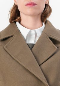Brown wool coat with a wide lapel, paired with a white button-up shirt and gray sweater, displaying a soft texture and minimalist design.