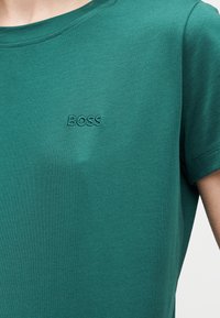 Πετρόλ κοντομάνικη μπλούζα με στρογγυλή λαιμόκοψη και μικρό κεντημένο λογότυπο "BOSS" στο στήθος, φορεμένη από ένα άτομο.