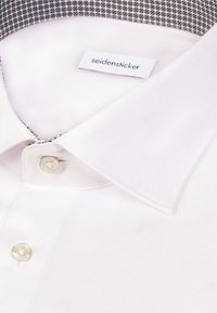 Biała koszula z kołnierzykiem wykonana z fakturowanego materiału, z czarno-białym wzorzystym wewnętrznym kołnierzykiem i zapięciem na pojedynczy guzik.