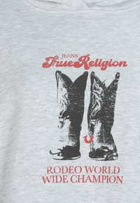 Para czarnych kowbojskich butów z nadrukiem czerwonego napisu "True Religion" oraz "Rodeo World Wide Champion" na jasnoszarej tkaninie.