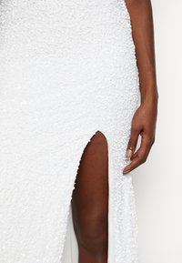 Vit paljettklänning med en hög slits; har en texturerad yta med små, runda paljetter och en figursydd silhuett. En hand syns vid sidan.