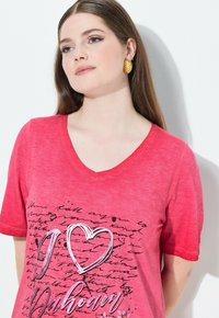 Camiseta de cuello en V de color rosa con un gráfico plateado "I ❤️ Ubahaan" sobre un patrón de estilo escrito a mano, hecha de una tela suave y texturizada.