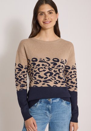 Femme souriante portant un pull beige et bleu marine à imprimé léopard et un jean bleu clair, debout devant un fond clair uni.
