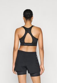 Sostén deportivo negro con diseño de espalda tipo racerback y detalle recortado, combinado con pantalones cortos deportivos negros sobre pantalones cortos ajustados de ciclismo.