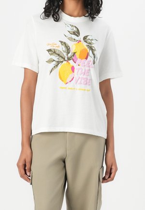 Biała koszulka z dużą grafiką dwóch cytryn i liści, różowy napis „Feel The Vibe”, noszona z beżowymi spodniami.