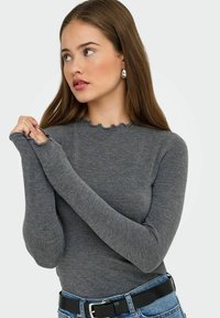 Sivý dlhý rukáv s okrajom s vlnkami. Tkanina vyzerá jemne a priliehavo, s predĺženými rukávmi, ktoré zakrývajú ruky. Skombinované s džínsami.