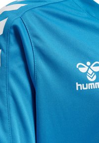 Turkis sportsjersey med glat overflade, med hvidt logo og designdetaljer på skuldrene. Korte ærmer, syntetisk stof.