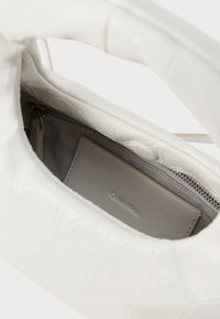 Geantă albă cu un model geometric embossat, un buzunar cu fermoar și un interior neted de culoare gri. Numele brandului este vizibil în interior.