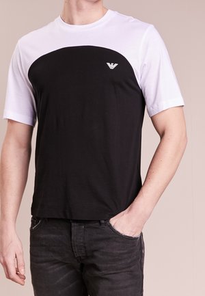 T-shirt a maniche corte bianco e nero con un pannello rotondo nero sul petto. Presenta un piccolo logo sul lato sinistro. Tessuto di texture liscia.