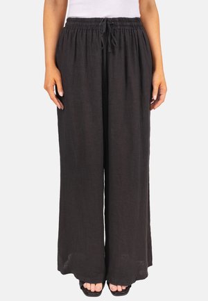 Femme portant un pantalon large noir à cordon de serrage ample, associé à des sandales noires à bout carré et un haut sans manches blanc.