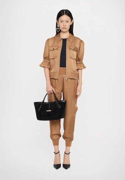 Bruine satijnen cropped jas met pofmouwen, bijpassende joggers, zwarte handtas met korte hengsels en zwarte puntige hakken.