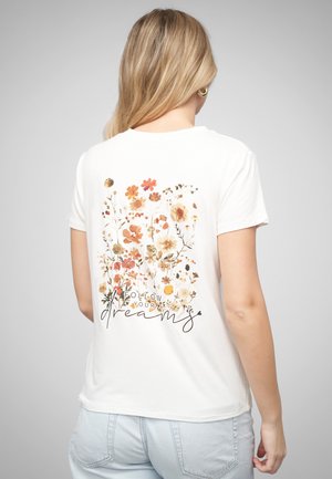 Lyshåret kvinde iført en hvid T-shirt med orange og beige blomsterprint og teksten "Følg dine drømme" på ryggen, kombineret med lyseblå jeans.