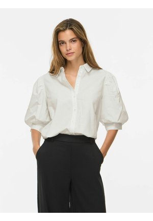 Femme portant une chemise blanche à manches bouffantes boutonnée, rentrée dans un pantalon noir taille haute, mains dans les poches, debout devant un fond uni.
