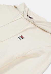 Krämfärgad hoodie i mjukt tyg. Har en dragsko i huvan, ribbad textur och en broderad logotyp med röda, blå och svarta detaljer.