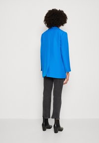 Personne aux cheveux bouclés portant un blazer bleu vif, un jean noir court et des bottines à talons noires, debout devant un fond uni.