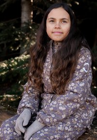 Paars gevoerd bloemenpak met een glanzende afwerking, voorzien van elastische manchetten en een hoge kraag. Grijze gebreide handschoenen zichtbaar. Natuurlijke achtergrond.