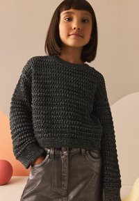 Pull noir en maille avec un motif texturé en relief, col rond et poignets côtelés, associé à un jean métallique gris foncé scintillant.