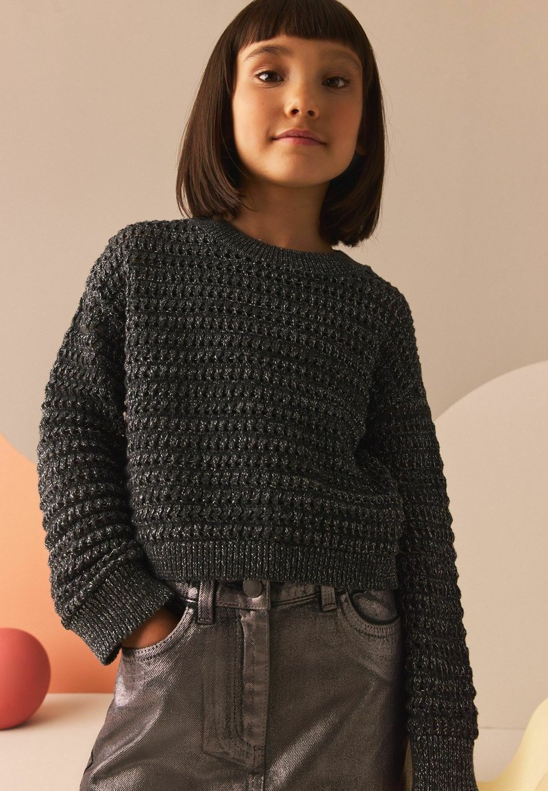 Pull noir en maille avec un motif texturé en relief, col rond et poignets côtelés, associé à un jean métallique gris foncé scintillant.