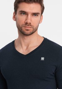 Mörkblå v-ringad långärmad tröja i mjukt material, med en liten vit logotyp på bröstet, figursydd design och slät textur.