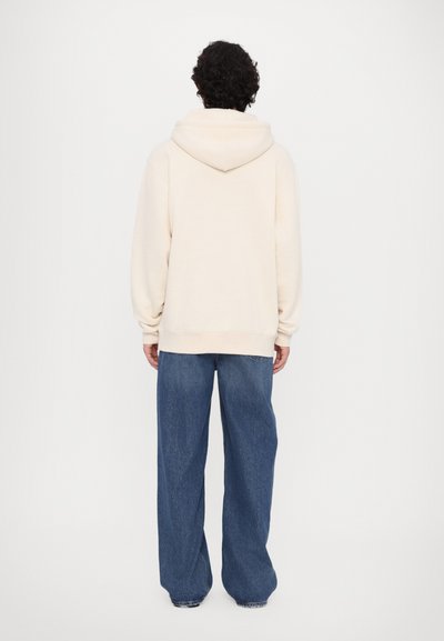 Cremfarbener Hoodie mit großer Kapuze, gerippten Bündchen und Saum, kombiniert mit lockeren blauen Jeans. Einfaches, minimalistisches Design.