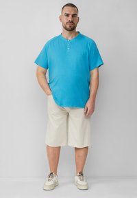 Koszula w turkusowym kolorze z krótkim rękawem, okrągłym dekoltem i zapięciem na guziki, połączona z beżowymi szortami do kolan i białymi sneakersami.