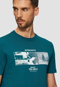 Teal puuvillapaita, jossa on graafinen printti, johon kuuluu kolme mustavalkoista kuvaa ja teksti "ARTS&CRAFTS" ylhäällä sekä "SERVICE 9302-309-0" alhaalla.