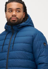 Blue puffer jacket with a zippered front, adjustable hood, and black accents. Features horizontal quilted design and a round logo patch on the sleeve.