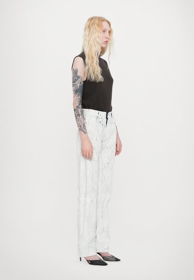 Black sleeveless top, white elastic waist pants with black paint splatter, and pointed black heels. Long wavy blonde hair, visible arm tattoo.