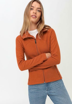 Oranje fleece zip-up jack met hoge kraag, nauwsluitend ontwerp, lange mouwen en zichtbare rits. Draagt over een lichtgekleurde shirt.