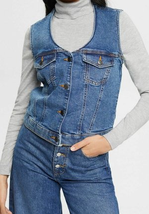 Persoon draagt een blauw denim mouwloos vest over een lichtgrijze coltrui en blauwe denim jeans met hand in de zak.