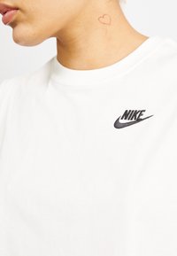 Camiseta blanca de algodón con un logotipo de Nike bordado en negro en el lado izquierdo del pecho y un pequeño tatuaje de corazón rojo en el cuello.