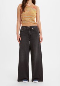 Woman wearing sleeveless multicolor knit top, high-waisted wide-leg black jeans, and blue sneakers, standing against a plain white background.