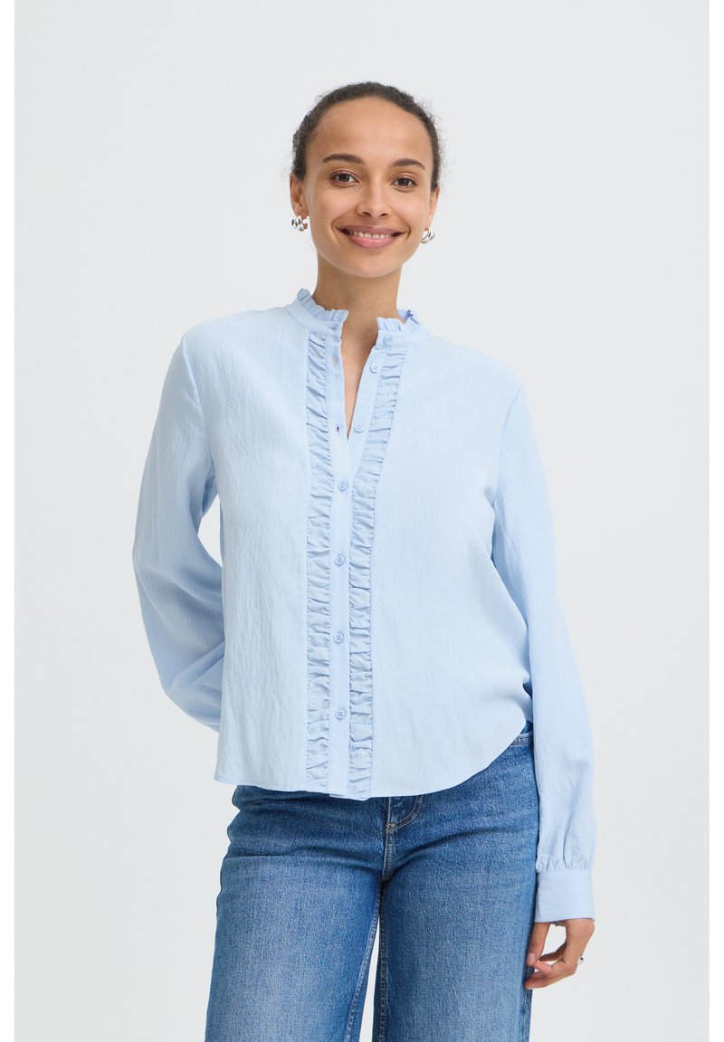 Femme souriante portant un chemisier bleu clair à volants boutonné et un jean bleu, debout avec une main dans le dos sur un fond blanc uni.