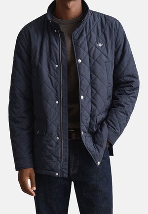 Homme portant une veste matelassée bleu marine avec des boutons-pression et une fermeture éclair, associée à un jean foncé et un pull marron en dessous.