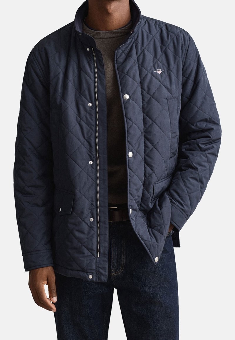 Homme portant une veste matelassée bleu marine avec des boutons-pression et une fermeture éclair, associée à un jean foncé et un pull marron en dessous.