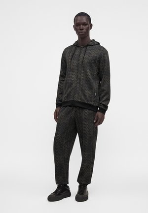 Homme portant un sweat à capuche et un pantalon sombres assortis avec un motif de texte clair répété, associé à des baskets noires, debout devant un fond uni.