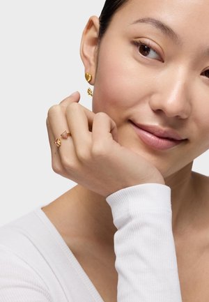 Młoda kobieta nosząca złote kolczyki w kształcie serca oraz złoty pierścionek z różowymi kamieniami, opierająca twarz na dłoni, delikatnie się uśmiechając.