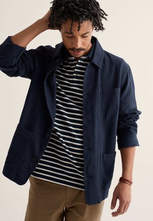 Mann mit lockigem Haar, der eine dunkelblaue Jacke über einem marineblau-weiß gestreiften Poloshirt trägt und mit einer Hand seine Haare berührt.